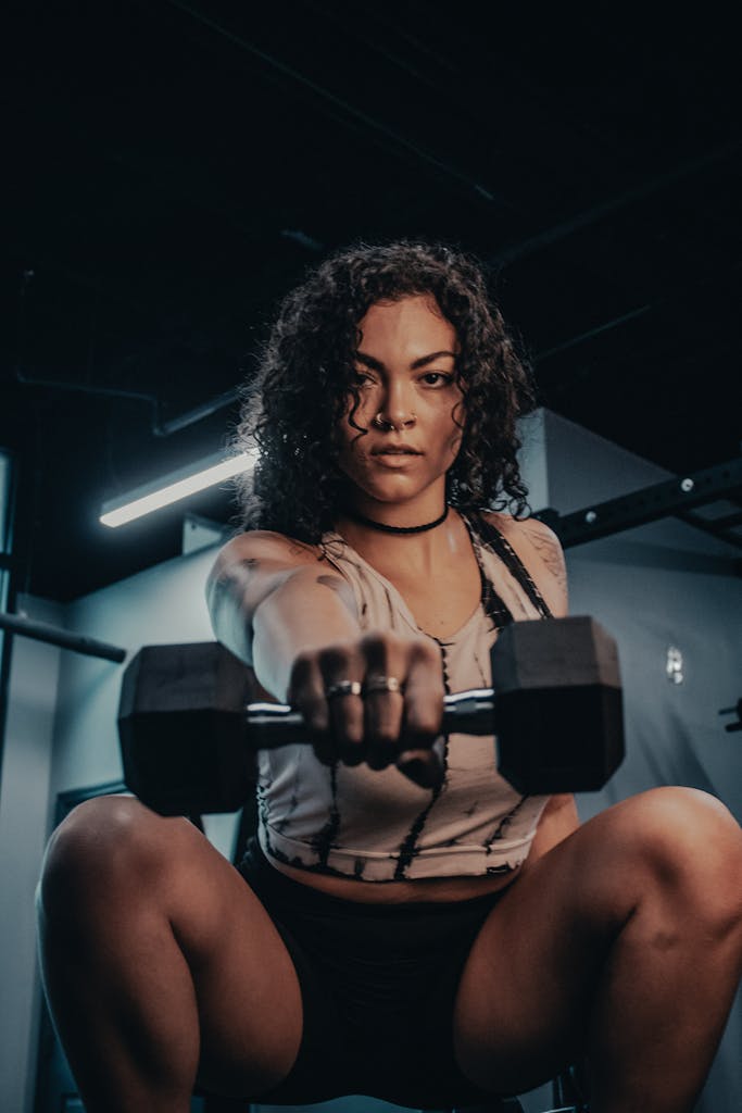 Focused woman with curly hair lifting a dumbbell in a dimly lit gym, showcasing strength.